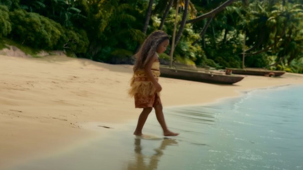 Young Moana standing at the shoreline as the ocean reaches toward in Moana Live Action.