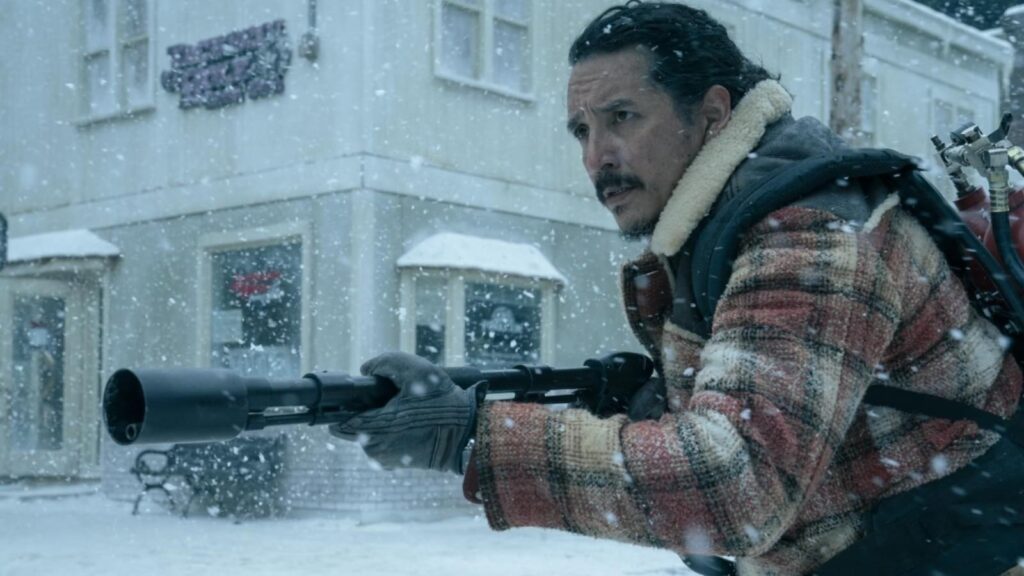 Gabriel Luna holding weapon in snowy street while wearing plaid winter jacket and backpack