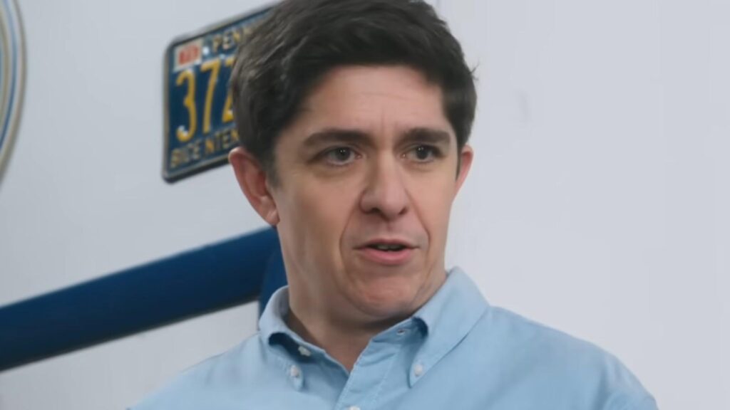 Steve Gerben as Will speaking inside tire shop office with vintage license plate backdrop in Tires Season 3