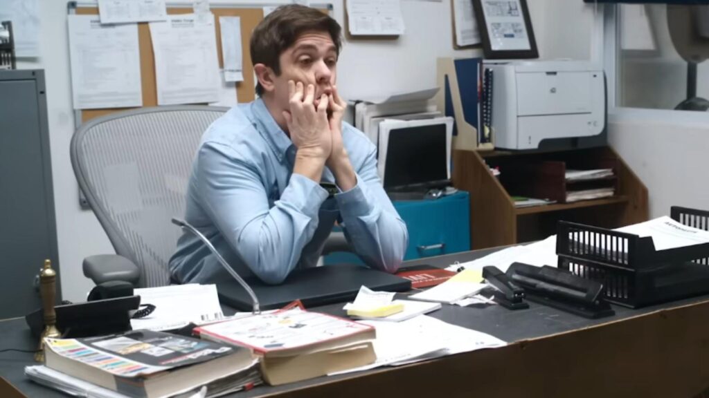 Shane Gillis as Shane sitting behind cluttered desk looking overwhelmed at work in Tires Season 3