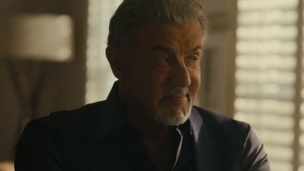 Sylvester Stallone with gray hair and beard wearing a dark suit indoors